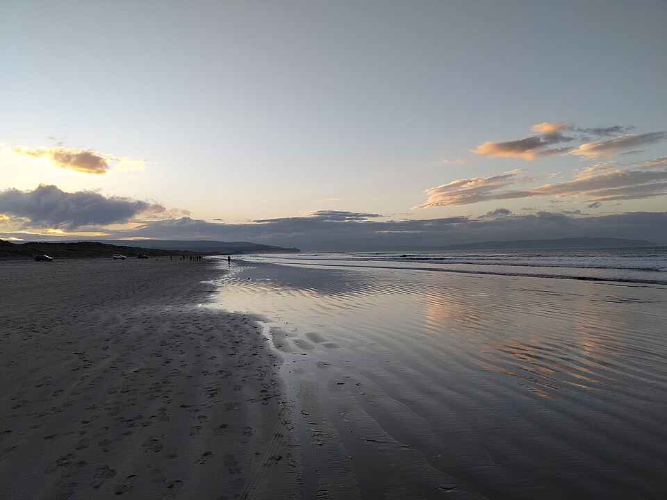 Wide sandy beach at Portstewart Strand with small waves and sunset reflections.