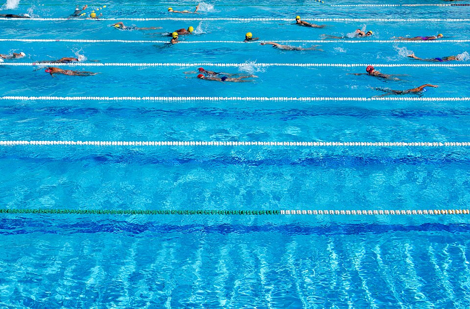 Swimmers move along marked outdoor pool lanes seen from above in Ilioupoli, Greece.