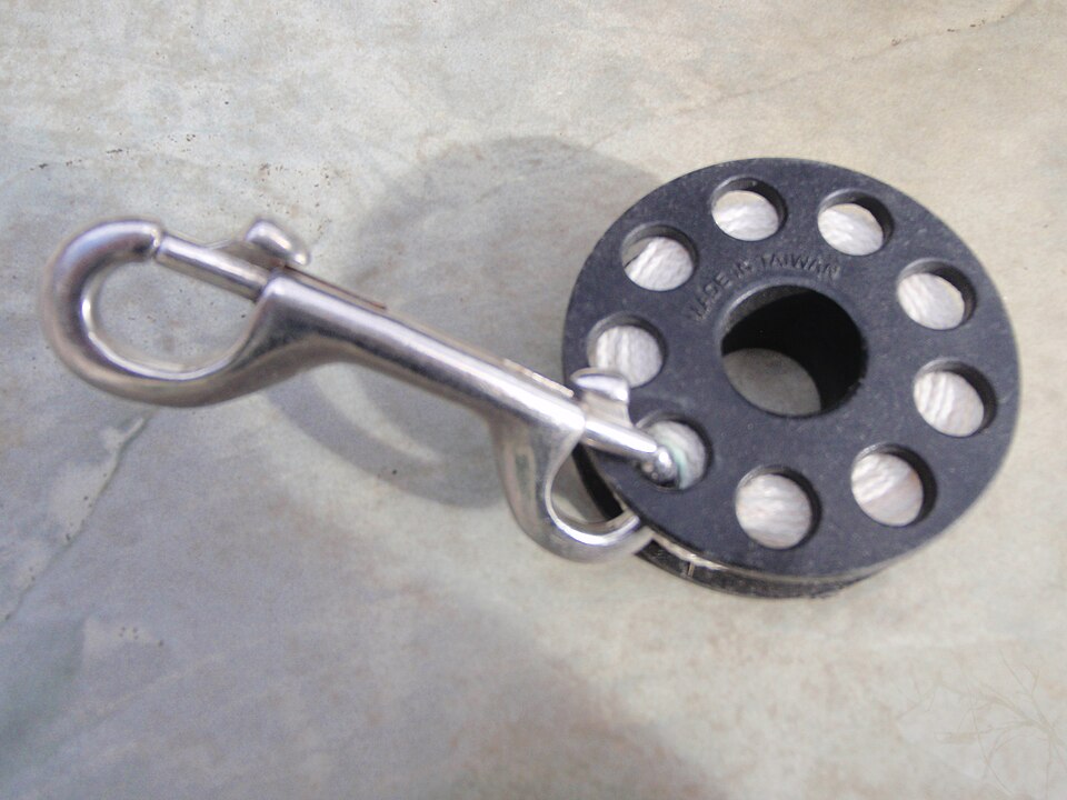 Archived photograph of a black plastic diving spool with a double-ended metal bolt snap on a plain surface.