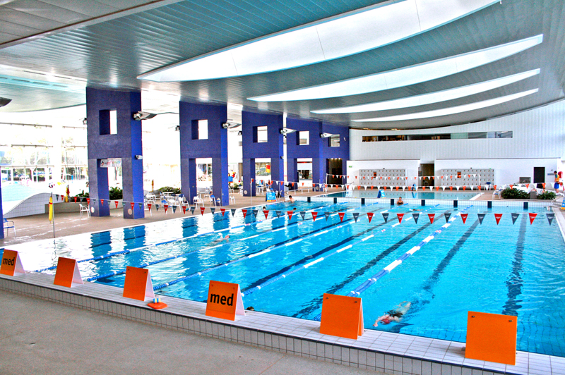 Indoor swimming pool with marked lanes, red and white flags, and swimmers in the water.