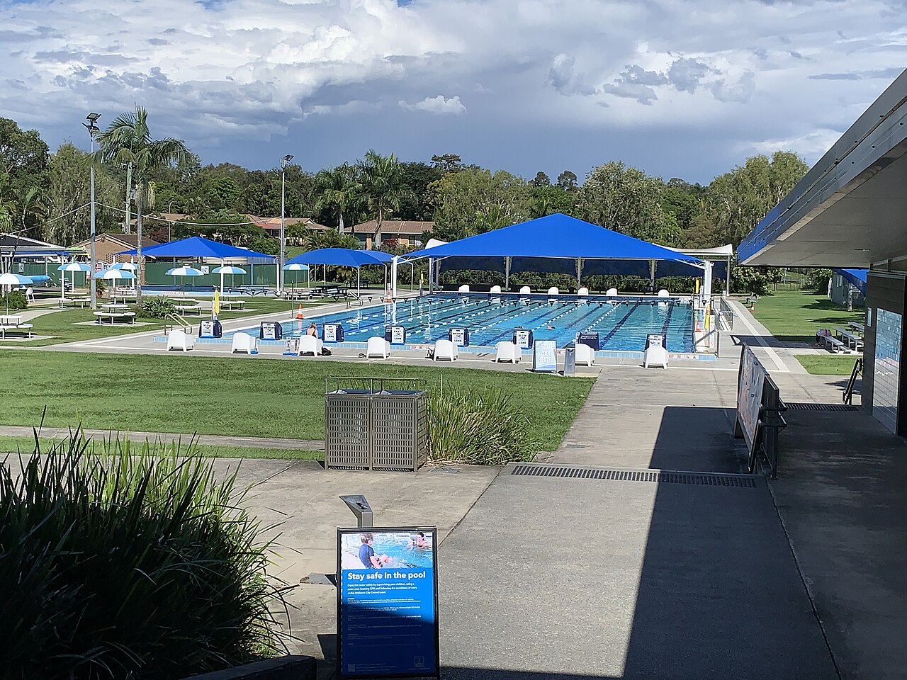 Outdoor swimming pool with marked lanes, shade sails and a pool safety notice.